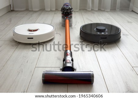 A black robot vacuum cleaner, a white robot vacuum cleaner, or a cordless vacuum cleaner with a nozzle lie on the laminate. Using modern technologies at home