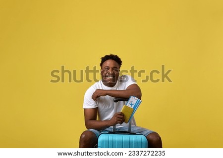 Happy traveler sat down to rest on a large suitcase, holding plane tickets in his hand. Positive African-American man posing in yellow studio with suitcase and documents in hand Stok fotoğraf © 