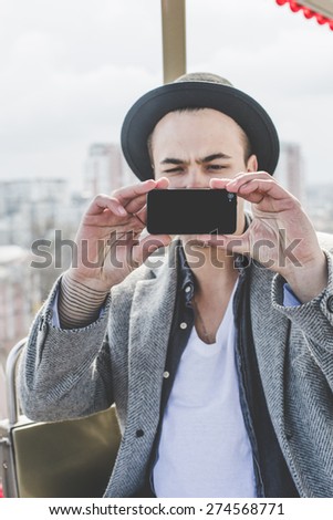 handsome young hipster guy in hat taking selfie with his smart phone in cabin of ferris wheel over city view from high background during sunny summer day on the festival