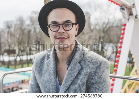handsome bearded young hipster guy in hat and glasses smiling and posing in cabin of ferris wheel over city view from high background during sunny summer day on the festival