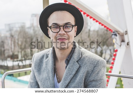handsome bearded young hipster guy in hat and glasses smiling and posing in cabin of ferris wheel over city view from high background during sunny summer day on the festival