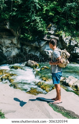 handsome man guy tourist standing alone by the mountain river waterfall forest during his summer vacation with his backpack