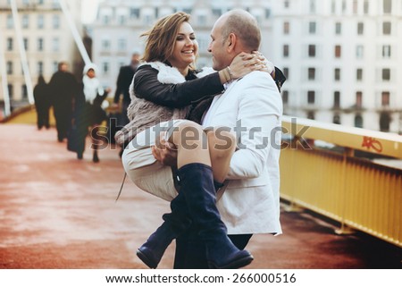 stylish beautiful happy groom holds the bride on hands walking on the bridge in Lyon cloudy