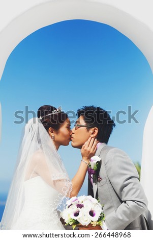 happy stylish asian bride and groom kissing under arch  on background blue sky in island Santorini