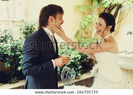 amazing beautiful rich stylish bride and groom smiling touching the face and drinking champagne goblet background wall and leaves