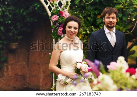 wonderful stylish rich happy bride and groom holding hands at a wedding ceremony smiling in green garden near white arch with flowers Rome Italy