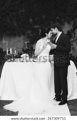 rich stylish happy bride and groom kissing  holding goblets with champagne  near a white wedding table decorated with flowers peonies and candles Rome Italy black and white