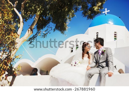happy stylish asian bride and groom sitting on a wall under green tree  on street of island Santorini background church and blue sky