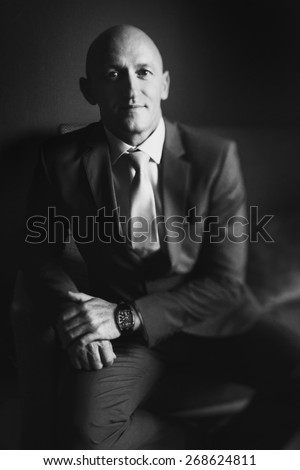 solid bald man in a suit sitting on a chair looking straight black and white
