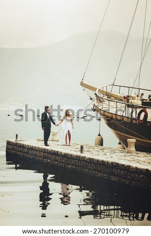 Stylish young bride and groom walking on the pier near yacht on background sea and mountains of Montenegro
