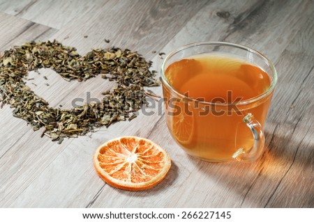Cup of tea standing on a wooden table. Next to a cup of heart made of dried tea leaves and round slice of dried orange
