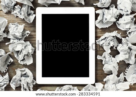 White tablet computer with a black screen on a wooden table. Around the tablet lies lot  crumpled paper