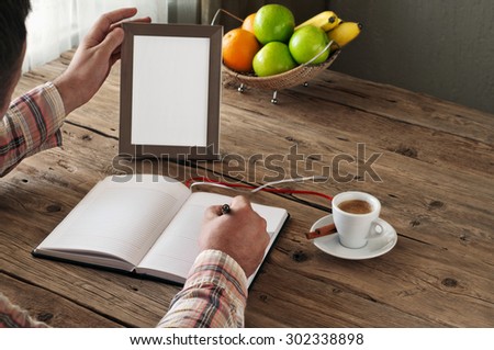 Hand of man writing in a blank notebook on the wooden table. Next on the table is an empty frame. Copy space. Free space for text. top view