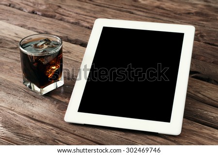 White tablet computer with a blank screen on the wooden table with a glass of cola with ice. Top view. Copy space. Free space for text.