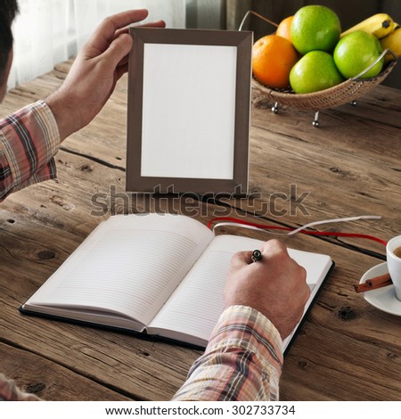 Hand of man writing in a blank notebook on the wooden table. Next on the table is an empty frame. Copy space. Free space for text. top view. Square frame