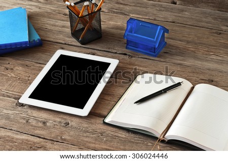 white tablet computer with a blank screen on wooden desk. Next to the tablet computer is on the table diary, pencils and piggy bank in the form of house. Copy space. Top view. Free space for text