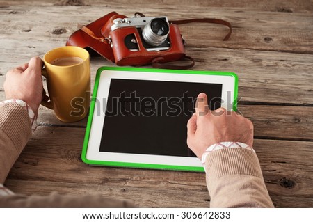 Male hand clicks then blank screen tablet computer on the wooden table and holding a cup of coffee closeup. Next on the table vintage camera. Copy space. Top view. Free space for text