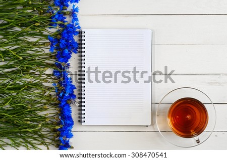White open notebook for notes on a white wooden table with a bouquet of blue flowers and a cup of tea. Top view. Copy space. Free space for text