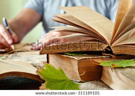 Old books. Old open book close up with autumn leaves on the table. In the background a man hand writing in a notebook. Copy space. Free space for text