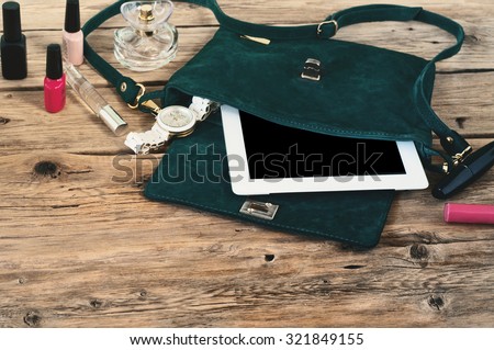 Suede female bag with tablet computer, watch and women cosmetics (lip gloss, mascara, nail polish and spirits)  on a wooden table closeup. Top view