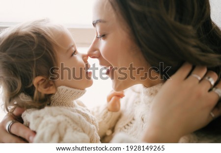 Young mother hugging her little daughter, they sitting near window and smiling. 