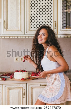 Young beautiful woman in an apron making cake with cream and berries at the kitchen