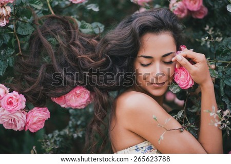 Beautiful young woman with long curly hair posing near roses in a garden