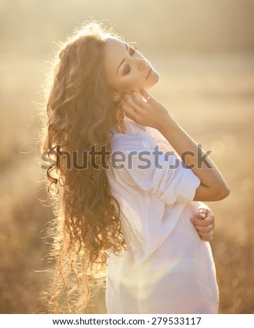 Young woman with beautiful long curly hair posing in a field at sunset. Summer mood