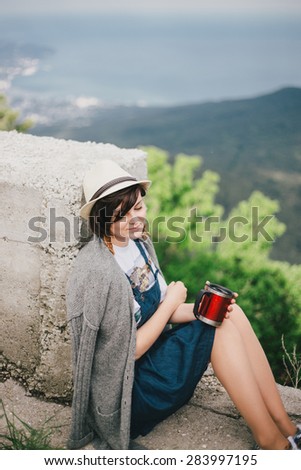 Young fashion woman sitting on a top of the mountain drinking tea with beautiful sea and hills view on the background