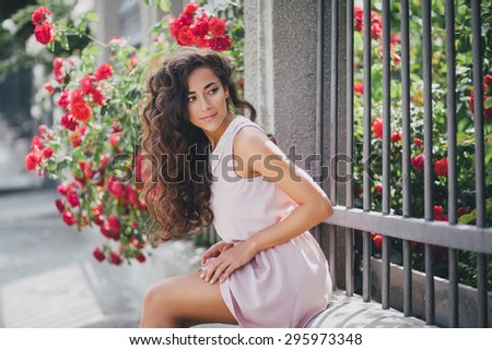 Beautiful young woman in a pink dress with long curly hair posing near roses in a garden