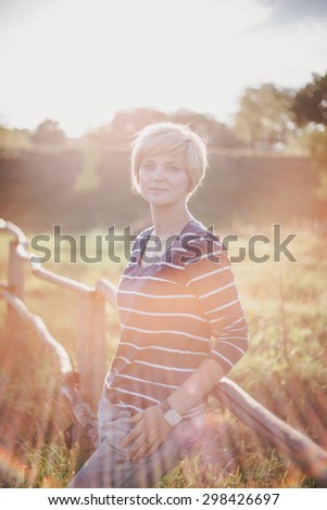 Young woman standing near fence on the country background at the sunset