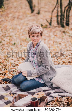 Young beautiful woman in a knitted sweater reading book and drinking tea in an autumn forest