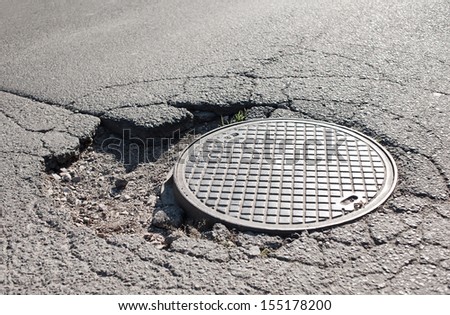 Large pothole in asphalt and circular manhole cover of sewage well in road, close metal sewer cover in Warsaw, Poland, horizontal orientation, nobody.