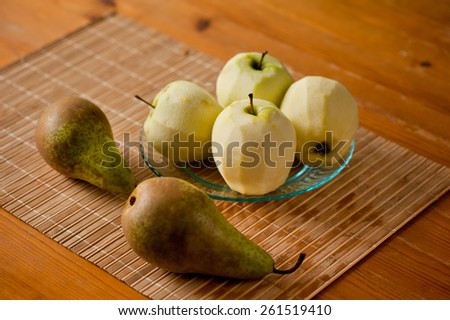 Peeled apples on glass plate and pears on bamboo mat, whole fruits on wooden table, nobody, horizontal orientation.