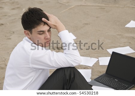 The young guy (businessman) sitting c and thinks on the beach. Around the guy scattered papers and laptop stands