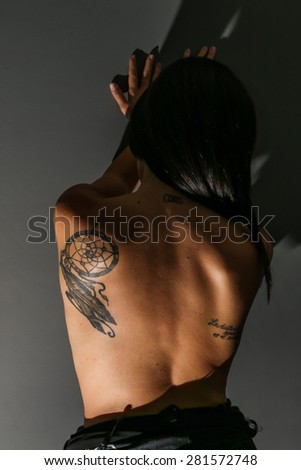 Model from behind, standing against a dark wall, showing her tattoo against envy people
