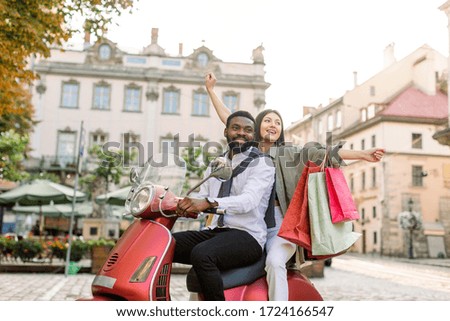 Cheerful glamorous young multiethnic couple riding red vintage scooter in the street, African man wears white shirt and pants and Caucasian woman holds colorful shopping bags with travel souvenirs