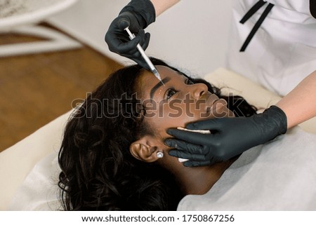 Close up of hands of doctor cosmetologist making the rejuvenating facial injections procedure for tightening and smoothing wrinkles on the face skin around the eyes for beautiful young African woman