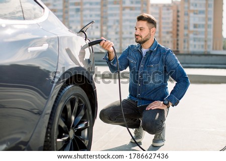 Hansome bearded guy sitting near his new modern electric car and holding plug of the charger, while car is charging at the charging station