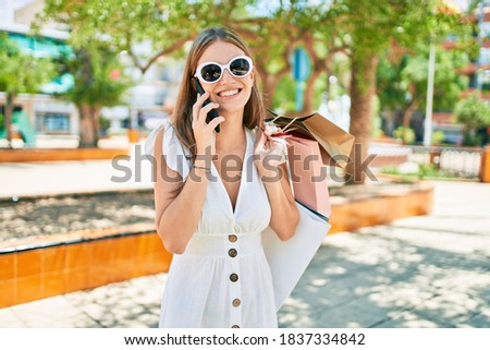 Young blonde woman smiling happy holding shopping bags and talking on the smartphone at street of city