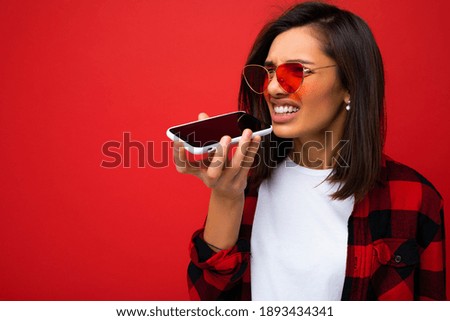 photo of beautiful angry young brunet woman wearing stylish red shirt white t-shirt and red sunglasses isolated over red background using mobile phone recording voice message looking to the side