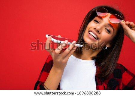 Photo shot of smiling attractive positive good looking young woman wearing casual stylish outfit poising isolated on background with empty space holding in hand and using mobile phone recording voice