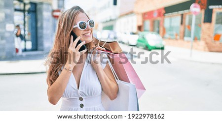 Young blonde woman smiling happy holding shopping bags and talking on the smartphone at street of city