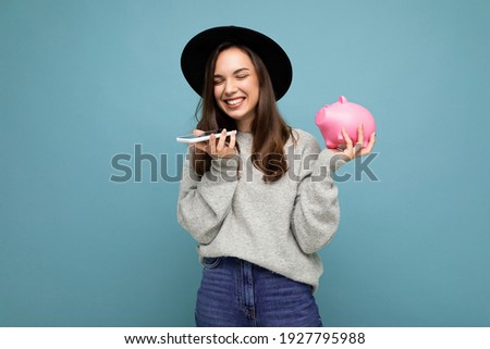 Photo of happy positive smiling laughing young beautiful brunette woman wearing stylish sweater and black hat isolated over blue background with empty space, holds pink piggy bank and uses mobile