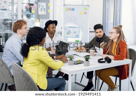 Group of five multiracial young people in casual outfit sitting at modern office and discussing new startup project. Creative students sharing with business ideas during meeting.
