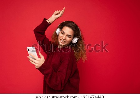 Shot of beautiful smiling young brunette curly woman wearing dark red sweater isolated over red background wall wearing white wireless headphones and listening to music and using mobile phone surfing