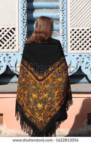 Russian girl in vintage traditional national Pavlovo Posad shawl and blue wooden house with carved windows, frames in Moscow city, Russia. Russian folk style in architecture and architectural fashion