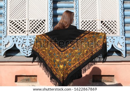 Russian girl in vintage traditional national Pavlovo Posad shawl and blue wooden house with carved windows, frames in Moscow city, Russia. Russian folk style in architecture and architectural fashion