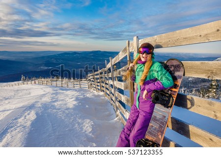 Successful young girl snowboarding in the mountains Sheregesh. Snowboarder resting.  Stylish caucasian snowboarder. Snowboarder smiling at the camera on top of the mountain on a background of sky. 