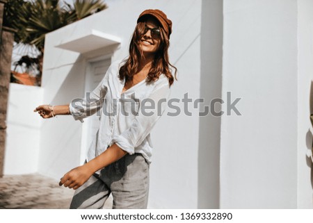 Pretty girl in summer blouse and linen pants walks outside. Portrait of woman in sunglasses and brown hat in yard of white building